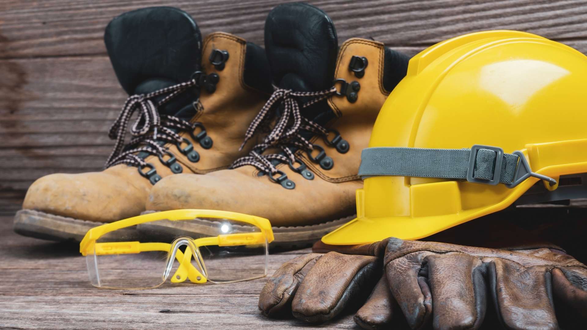 Foto con scarpe antinfortunistiche marroni, occhiali protettivi gialli, caschetto giallo e guanti grossi da lavoro marroni