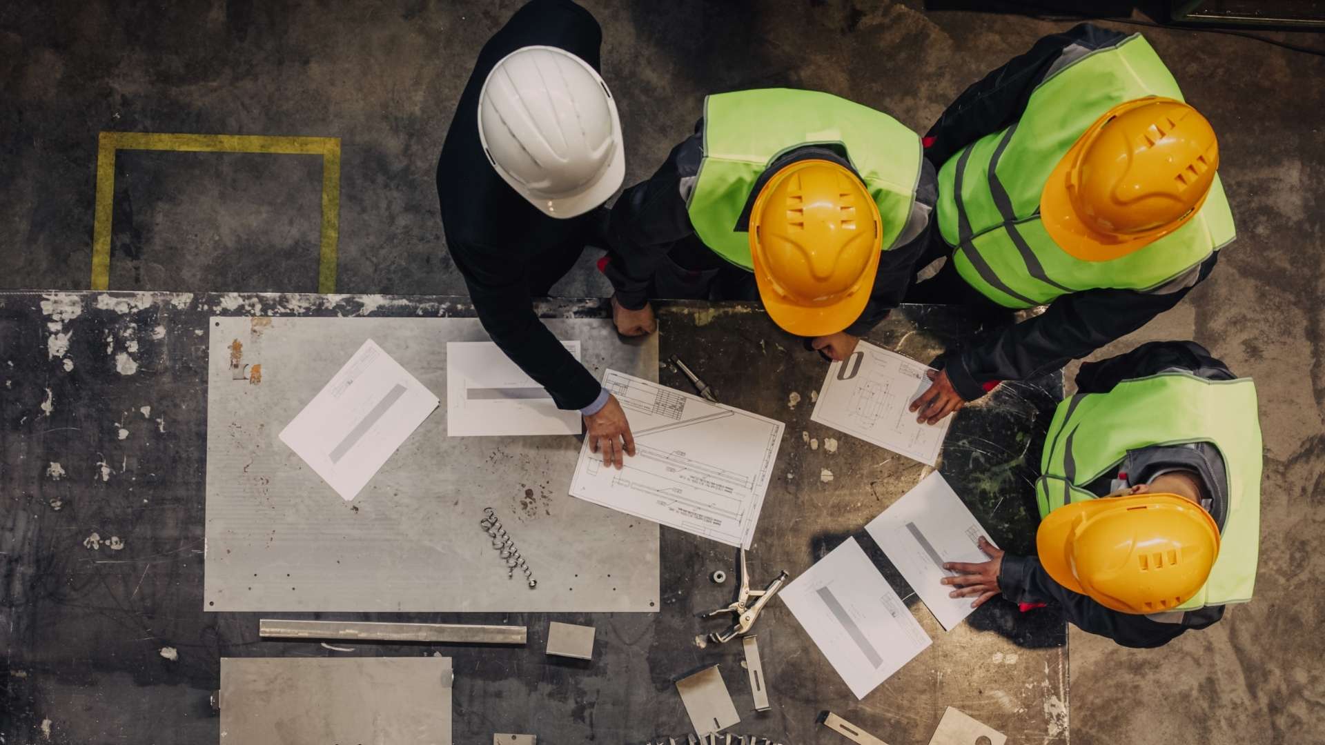 Quattro uomini con elmetto e giubetto trivalente fotografati dall'alto mentre guardano progetti su un tavolo da lavoro.