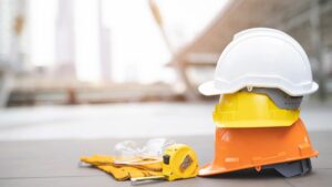 Tre elmetti uno sopra l'altro (arancione, giallo e bianco) appoggiati a terra con affianco un paio di guanti da lavoro, un metro e occhiali di protezione