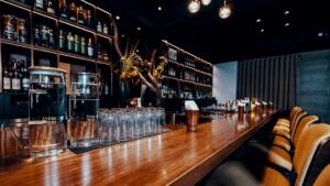 Bancone di legno di un bar, con sedie a sgabello da un lato e scaffale con bottiglie e vari materiali da bar