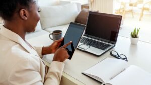Donna di colore, seduta al tavolo a casa, con il divano sullo sfondo che lavora in smart working. Sul tavolo sono presenti il PC, l'agenda, gli occhiali, una tazza e un portapenne. In mano regge un tablet.