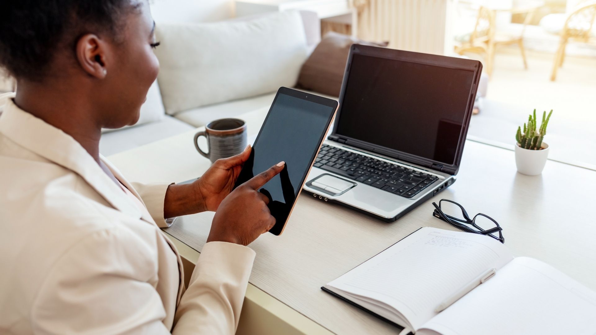 Donna di colore, seduta al tavolo a casa, con il divano sullo sfondo che lavora in smart working. Sul tavolo sono presenti il PC, l'agenda, gli occhiali, una tazza e un portapenne. In mano regge un tablet.